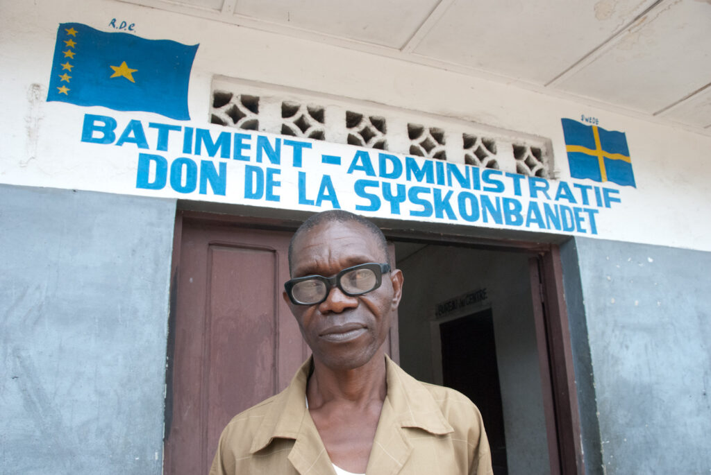 Afrikansk man står utanför byggnad där det ovanför dörren står "Administrativa byggnaden - gåva från Syskonbandet" på franska.